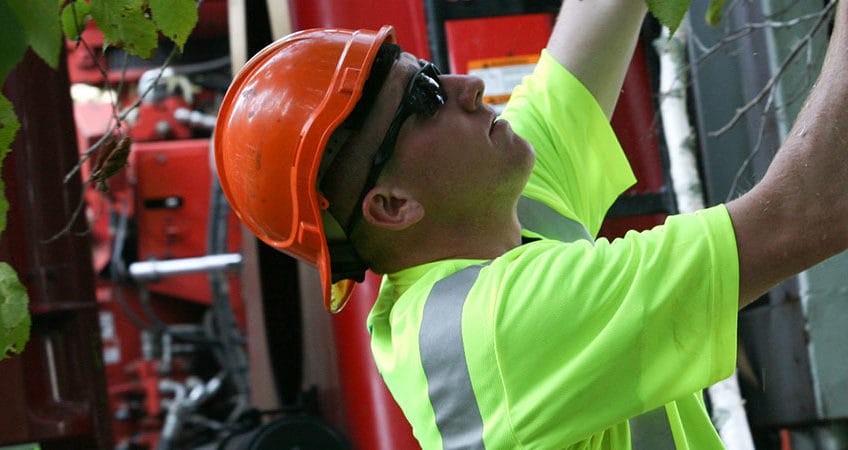 Tree service worker in a reflective safety shirt and orange hard hat using a chainsaw to cut branches near a wood chipper.